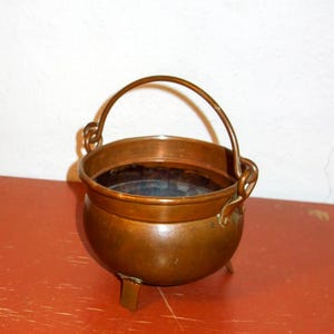 May include: A small, antique copper cauldron with a rounded body, three short legs, and a curved handle. The cauldron sits on a red wooden surface against a white wall. The copper has a warm, aged patina.