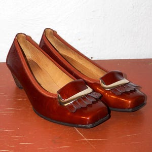 Puede incluir: Un par de zapatos vintage de cuero brillante, de color marrón rojizo, con tacón bajo. Cada zapato presenta una puntera cuadrada, una solapa decorativa con un acento dorado y un pequeño detalle de flecos. Los zapatos se exhiben sobre una superficie marrón rojizo.