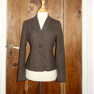 May include: A brown blazer with a tailored fit, displayed on a mannequin. The jacket has a notched collar, two front pockets, and button closures. The mannequin is on a metal stand, set against a wooden door.