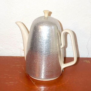 以下が含まれることがあります： クリーム色の取っ手、注ぎ口、蓋が付いた、ヴィンテージの銀色のティーポット。 ティーポットはハンマー加工された質感で、赤茶色の表面に置かれています。 背景は白い壁です。