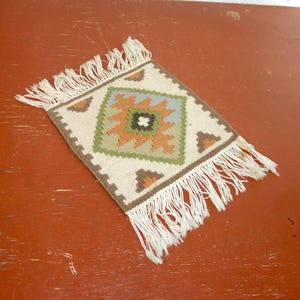 May include: A small, square woven rug with a geometric design. The rug features a central diamond pattern in green, orange, and blue, surrounded by brown and cream-colored borders. White fringe edges the rug.