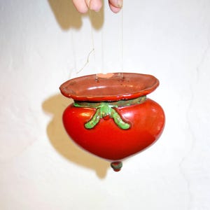 May include: A heart-shaped, red ceramic ornament with a brown lid and green accents. The ornament is suspended by a thin string, suggesting it's a hanging decoration. The design is simple and charming.