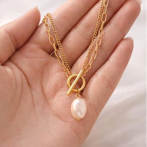 Könnte beinhalten: Goldfarbene Halskette mit Doppelketten-Design und Knebelverschluss. Die Halskette hat einen hängenden, oval geformten Perlenanhänger. Der Text "THE SOULBABE STUDIO" und "Jewelry" sind sichtbar.