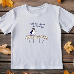 May include: White t-shirt featuring a cartoon penguin and the text "I used to be indecisive. Now I'm not sure." The penguin stands near signs that say "Yes?", "No?", "Maybe?", and "Ask again?"