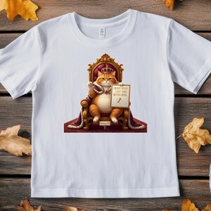 May include: White t-shirt featuring a cartoon cat wearing a crown and sitting on a throne. The cat holds a sign that reads "I'M NOT BOSSY, I JUST HAVE BETTER IDEAS." The design includes gold and red accents.