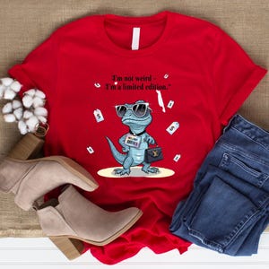 May include: A red t-shirt featuring a cartoon lizard wearing sunglasses and holding a purse. The text on the shirt reads, "I'm not weird - I'm a limited edition." The shirt is paired with jeans and beige ankle boots.