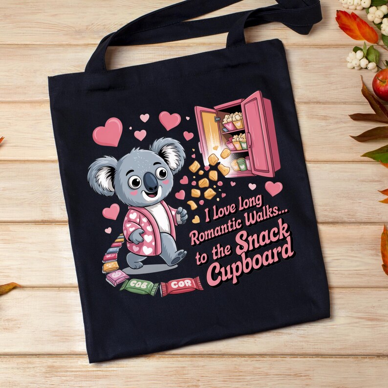 May include: Black tote bag featuring a cartoon koala wearing a pink jacket with hearts. The koala is walking towards a pink snack cupboard. Text reads "I love long romantic walks... to the Snack Cupboard."