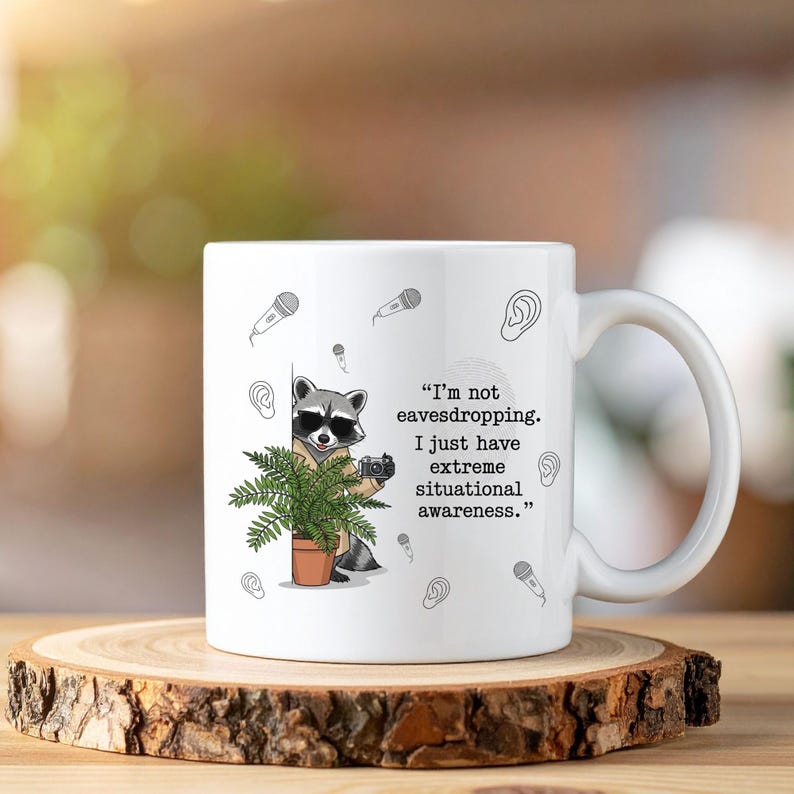 May include: White ceramic mug featuring a raccoon wearing sunglasses and holding a camera, with a potted plant. The mug has the text: "I'm not eavesdropping. I just have extreme situational awareness." Decorative microphones and ears are also printed on the mug.