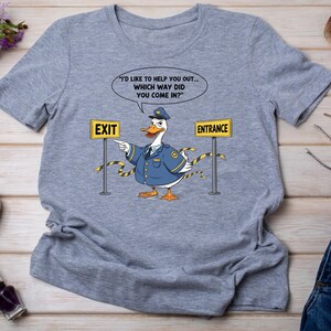 May include: A light gray t-shirt featuring a cartoon duck dressed as a police officer. The duck points towards an "EXIT" sign and has a speech bubble with the text "I'D LIKE TO HELP YOU OUT... WHICH WAY DID YOU COME IN?" and an "ENTRANCE" sign.