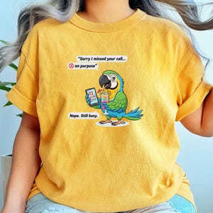 May include: Mustard yellow t-shirt featuring a cartoon parrot holding a cocktail and a phone with 12 missed calls. The parrot has blue, green, and yellow feathers. Text on the shirt reads, "Sorry I missed your call... on purpose" and "Nope. Still busy."