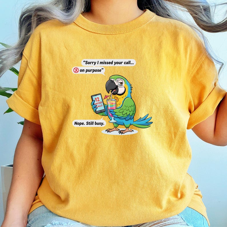 May include: Mustard yellow t-shirt featuring a cartoon parrot holding a cocktail and a phone with 12 missed calls. The parrot has blue, green, and yellow feathers. Text on the shirt reads, "Sorry I missed your call... on purpose" and "Nope. Still busy."