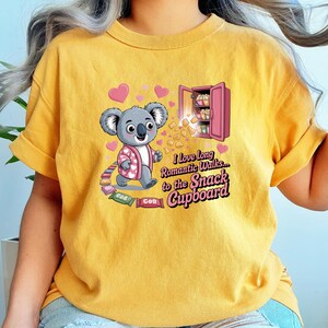 May include: Mustard yellow t-shirt featuring a cartoon koala wearing a pink robe with a heart pattern. The koala walks towards a pink snack cupboard. Text reads "I love long romantic walks to the Snack Cupboard."
