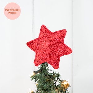 May include: A red, crocheted star tree topper with a glittery finish, placed atop a small Christmas tree. The star is a five-pointed design with a textured surface. The text "PDF Crochet Pattern" is in a pink circle in the upper left corner.