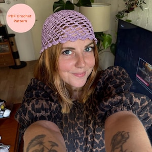 Op de afbeelding: Een lichtpaarse gehaakte muts met een opengewerkt netpatroon en een geschulpte rand. De muts heeft een cirkelvormige tekstoverlay in de linkerbovenhoek met de tekst PDF Crochet Pattern in oranje letters op een lichtroze achtergrond.