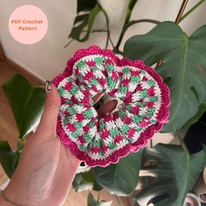 Puede incluir: Una scrunchie de ganchillo hecha a mano con un vibrante patrón de rosa, blanco y verde. La scrunchie presenta un borde festoneado y se sostiene frente a una planta verde frondosa. El texto en la imagen dice "PDF Crochet Pattern."