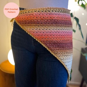 Op de afbeelding: Een gehaakte omslagdoek in tinten beige, perzik, oranje en bordeaux. De omslagdoek heeft een driehoekige vorm met een geschulpte rand en wordt gedragen over een donkerblauwe spijkerbroek. De tekst "PDF Crochet Pattern" staat in een roze cirkel linksboven.