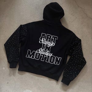 May include: Black hooded sweatshirt with white text that reads "ART Samyo IN Studios MOTION". The sleeves are dotted with small, shiny embellishments. The sweatshirt is on a gray surface.