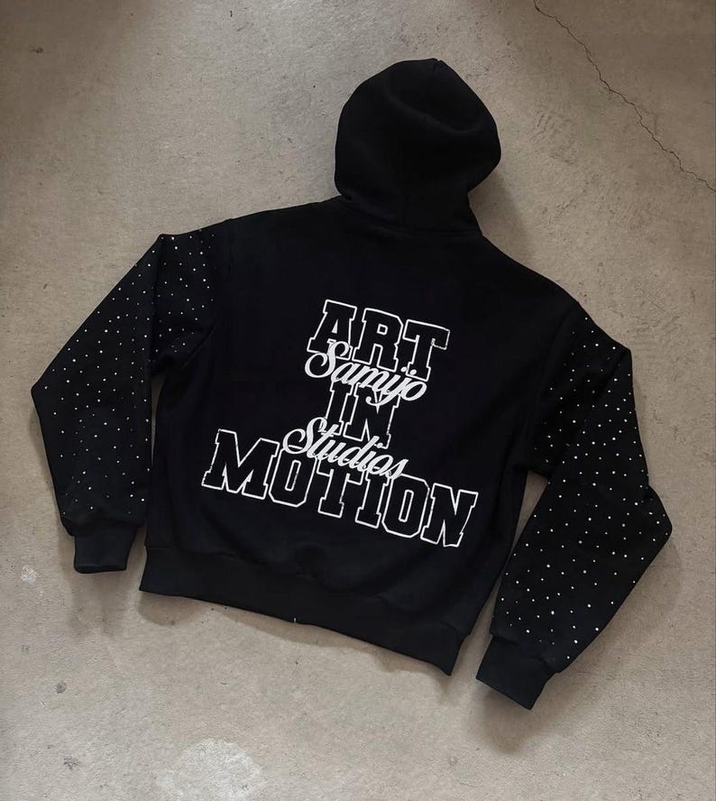 May include: Black hooded sweatshirt with white text that reads "ART Samyo IN Studios MOTION". The sleeves are dotted with small, shiny embellishments. The sweatshirt is on a gray surface.
