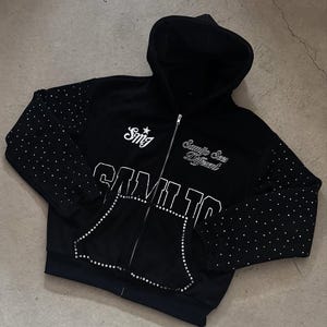 May include: Black zip-up hoodie with white text and rhinestone embellishments. The hoodie features the text "Samilto" and "Samejo Sees Different". Rhinestones are scattered on the sleeves and outline the pockets.