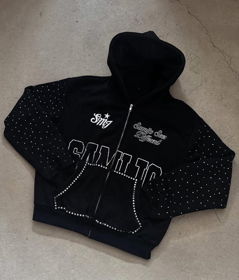 May include: Black zip-up hoodie with white text and rhinestone embellishments. The hoodie features the text "Samilto" and "Samejo Sees Different". Rhinestones are scattered on the sleeves and outline the pockets.