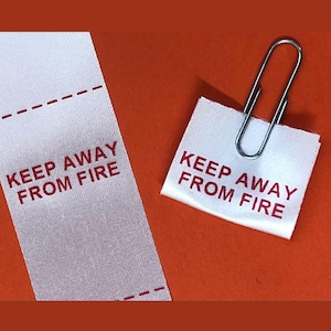 May include: Two white fabric labels with red text reading "KEEP AWAY FROM FIRE" are shown against an orange backdrop. One is a strip with a dashed line, the other a small square secured by a silver paper clip. Safety label.
