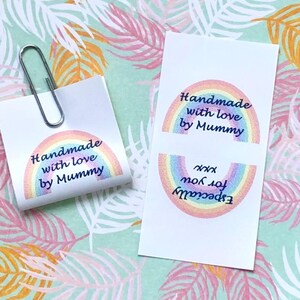 Op de afbeelding: Twee rechthoekige witte papieren labels met een kleurrijke regenboogafbeelding en de tekst "Handmade with love by Mummy" en "Especially for you xxx". E&eacute;n label is gevouwen en vastgezet met een zilveren paperclip, de andere ligt plat.