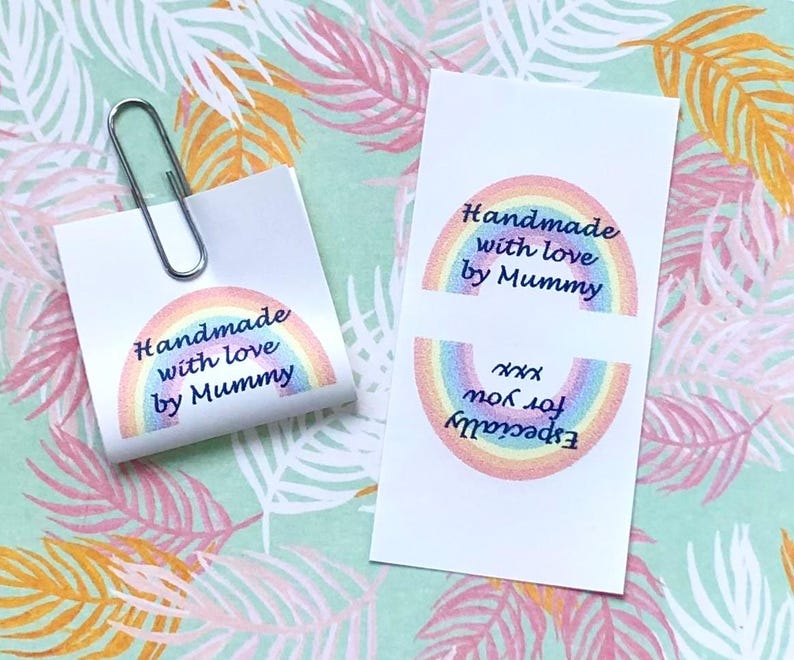 Op de afbeelding: Twee rechthoekige witte papieren labels met een kleurrijke regenboogafbeelding en de tekst "Handmade with love by Mummy" en "Especially for you xxx". E&eacute;n label is gevouwen en vastgezet met een zilveren paperclip, de andere ligt plat.