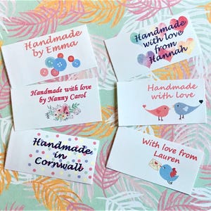 May include: Several white rectangular labels with handwritten text and illustrations. The labels read phrases such as "Handmade by Emma," "Handmade with love from Hannah," and "Handmade in Cornwall." Designs include buttons, hearts, birds, and floral arrangements.