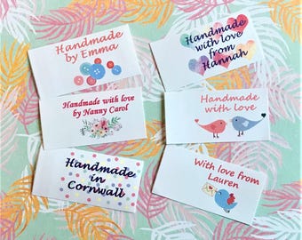 Op de afbeelding: Zes rechthoekige witte stoffen labels met diverse ontwerpen en teksten. De labels bevatten: Handmade by Emma met knopen, Handmade with love from Hannah met hartjes, Handmade with love by Nanny Carol met bloemen, Handmade with love met vogels, Handmade in Cornwall met stippen, en With love from Lauren met een vogel en een envelop.