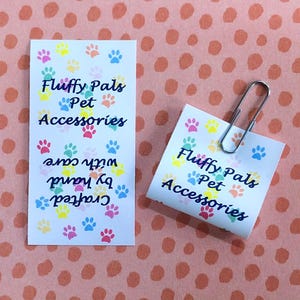 Op de afbeelding: Twee witte rechthoekige papieren labels met een kleurrijk patroon van kleine pootafdrukken in roze, geel, blauw en groen. De tekst luidt Fluffy Pals Pet Accessories en Crafted by hand with care. Een zilveren metalen paperclip is aan &eacute;&eacute;n label bevestigd.
