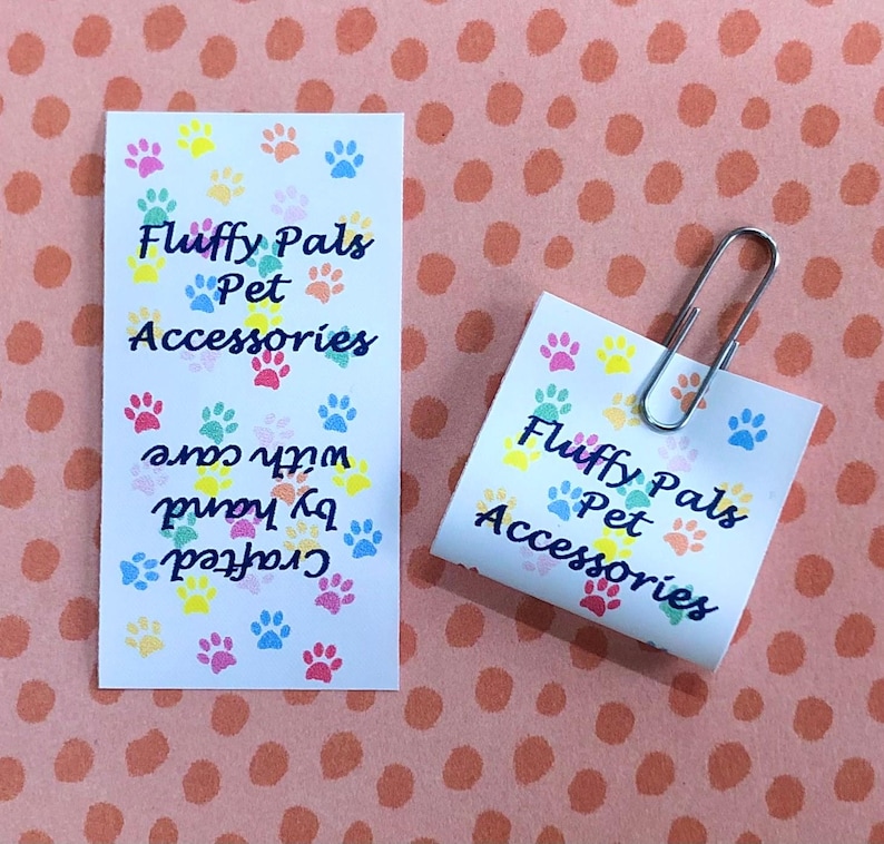 Op de afbeelding: Twee witte rechthoekige papieren labels met een kleurrijk patroon van kleine pootafdrukken in roze, geel, blauw en groen. De tekst luidt Fluffy Pals Pet Accessories en Crafted by hand with care. Een zilveren metalen paperclip is aan &eacute;&eacute;n label bevestigd.