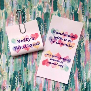 Op de afbeelding: Twee witte rechthoekige papieren labels met aquarelachtige hartjes in pasteltinten. E&eacute;n vierkant label met een zilverkleurige metalen paperclip bevat de tekst Betty's Boutique. Het andere label bevat de tekst Handmade with love by Lucinda en For you to treasure forever and ever.