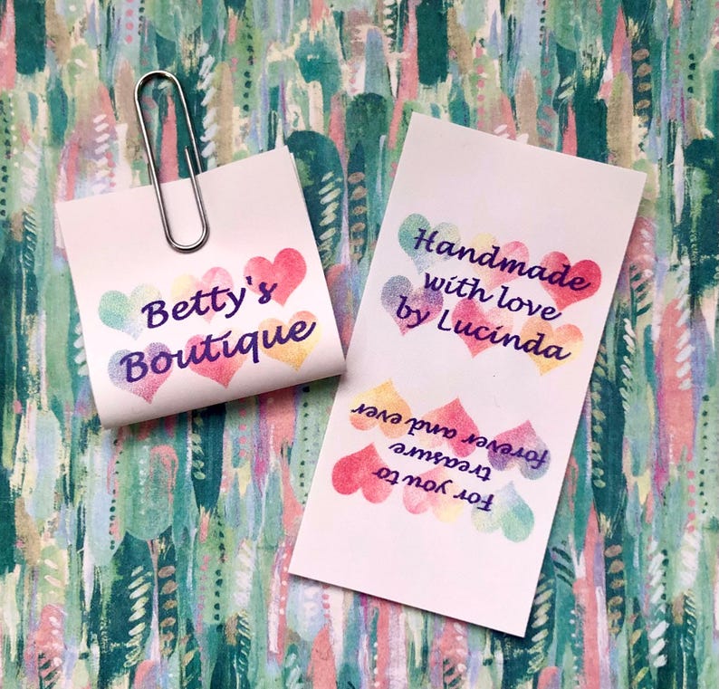 Op de afbeelding: Twee witte rechthoekige papieren labels met aquarelachtige hartjes in pasteltinten. E&eacute;n vierkant label met een zilverkleurige metalen paperclip bevat de tekst Betty's Boutique. Het andere label bevat de tekst Handmade with love by Lucinda en For you to treasure forever and ever.