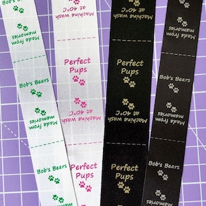 Op de afbeelding: Vier stoffen labels met tekst en pootafdrukken. De labels zijn wit, zwart en met patronen. De tekst bevat "Bob's Bears", "Perfect Pups", "Made from memories" en "Machine wash at 40°C". De pootafdrukken zijn groen, roze en goud.