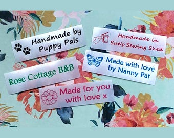Op de afbeelding: Een verzameling witte stoffen labels met verschillende teksten en afbeeldingen. De labels bevatten zinnen als "Handmade by Puppy Pals", "Rose Cottage B&B" en "Made with love". De achtergrond heeft een bloemenpatroon.