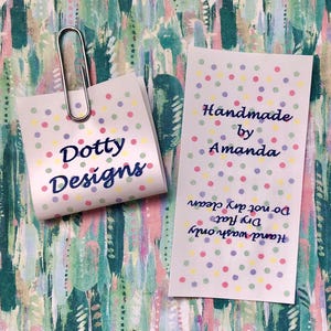Op de afbeelding: Twee witte stoffen labels met een kleurrijk stippenpatroon. Een vierkant label wordt vastgehouden door een zilveren metalen paperclip en toont de tekst Dotty Designs. Het rechthoekige label vermeldt Handmade by Amanda, Hand wash only, Dry flat, Do not dry clean.