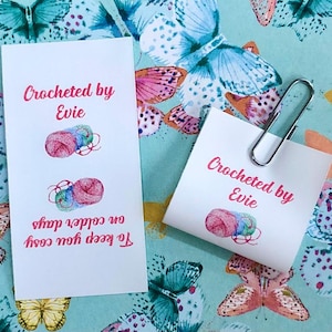 Op de afbeelding: Twee witte rechthoekige papieren labels met de roze tekst Crocheted by Evie en illustraties van kleurrijke bollen wol. E&eacute;n label bevat de extra roze tekst To keep you cosy on colder days en er is een zilveren metalen paperclip bevestigd.