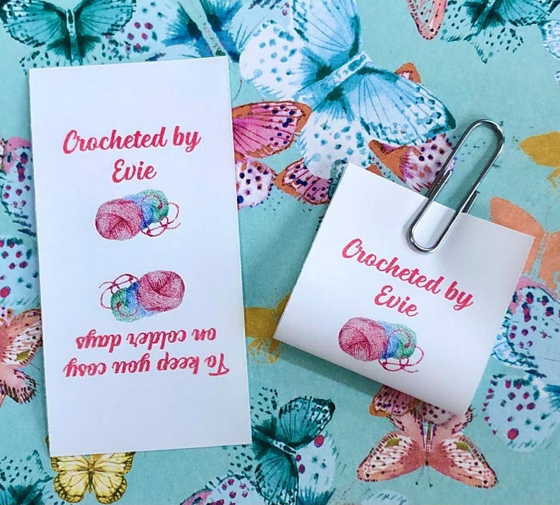 Op de afbeelding: Twee witte rechthoekige papieren labels met de roze tekst Crocheted by Evie en illustraties van kleurrijke bollen wol. E&eacute;n label bevat de extra roze tekst To keep you cosy on colder days en er is een zilveren metalen paperclip bevestigd.