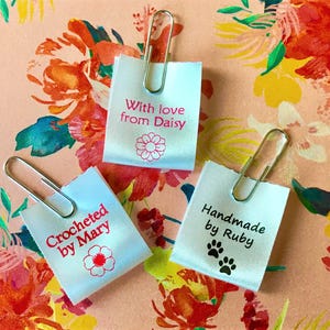 May include: Three small, white fabric tags with text and paper clips. One says "With love from Daisy" with a flower graphic. Another says "Crocheted by Mary" with a flower. The third says "Handmade by Ruby" with paw prints. The background is a floral pattern.