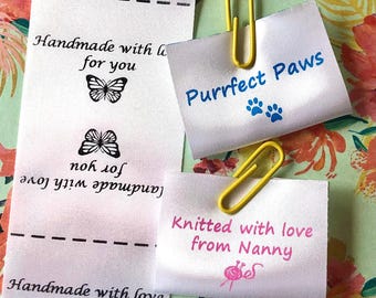 Pode incluir: Etiquetas de tecido branco com texto e gráficos. Uma etiqueta diz "Handmade with love for you" com ilustrações de borboletas. Outra diz "Purrfect Paws" com pegadas. Uma terceira diz "Knitted with love from Nanny".