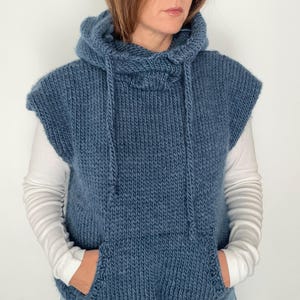Könnte beinhalten: Eine blaue Kapuzenweste aus Strick mit einer Kängurutasche. Die ärmellose Weste hat einen Kordelzug an der Kapuze. Die Person trägt ein weißes Langarmshirt und eine blaue Jeans. Das Kleidungsstück ist aus dickem Garn gefertigt.