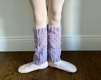 May include: A pair of lavender knitted leg warmers with a diamond pattern. The leg warmers are worn over white tights and ballet shoes.