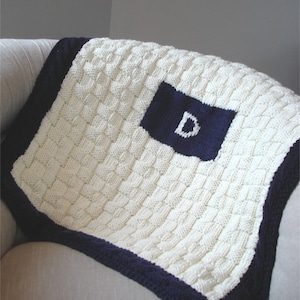 Pode incluir: Cobertor de malha creme com uma borda azul marinho e um quadrado azul marinho com a letra "D". O cobertor tem um padrão texturizado e tecido. O cobertor está dobrado sobre uma superfície de cor clara.