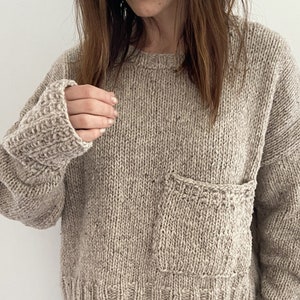 以下が含まれることがあります： 前面に大きなパッチポケットが付いた、茶色のニットセーター。ゆったりとしたフィット感とクルーネックです。