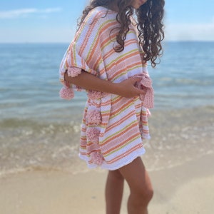 May include: A white and pink striped knit cover-up with pink pom-poms around the edges. The cover-up is worn by a person standing on a sandy beach.