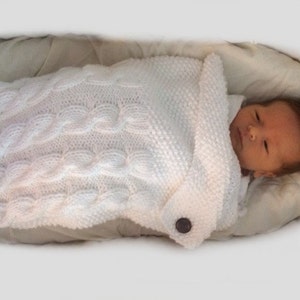 Könnte beinhalten: Eine weiße gestrickte Babydecke mit einem Zopfmuster und einem Knopfverschluss. Ein Baby schläft friedlich in der Decke.