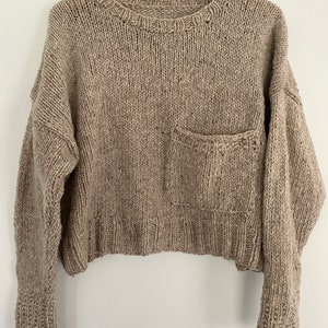 以下が含まれることがあります： 前面にパッチポケットが付いたベージュのニットセーター。長袖でゆったりとしたフィット感です。