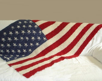 May include: A knitted American flag blanket draped over a white couch. The blanket is made of red, white, and blue yarn and features a traditional American flag design.