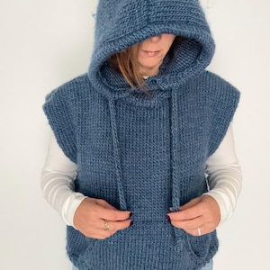 Könnte beinhalten: Eine blaue Kapuzenweste aus Strick mit Kordelzug. Die Weste hat eine Kängurutasche und ist ärmellos. Die Person auf dem Bild trägt ein weißes Langarmshirt und eine blaue Jeans.