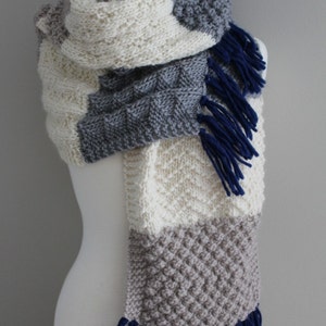 May include: A knitted scarf with a white, gray, and beige color scheme. The scarf has a variety of knit patterns and blue tassels on the ends.  PDF Pattern.